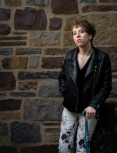Woman with short brownish curly stands with her cane by a cobblestone brick wall. She is wearing a leather jacket and white print pants.