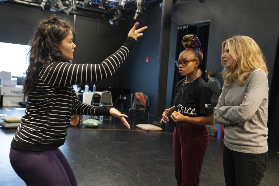 Three women actively confer in a rehearsal studio; the first, at left, has dark, wavy hair and is wearing a black-and-white striped top, and has her arms raised in a theatrical gesture; the other two, one a young woman in a black T-shirt with dark hair piled up in a bun, the other a woman in a grey top with long blond hair, look at the first woman in listening and thinking poses.