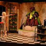 A production image in which a blonde white leading lady sings, her arms outstretched, on the set of a flower shop; behind her to left is a white man dressed as a giant plant, and far stage left, a bespectacled young white man, looking nervously, holds up a telephone receiver.