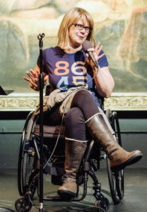 A blond woman in a wheelchair speaks into a mic at a public presentation; she is wearing an 86 45 T-shirt.