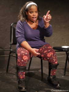 A Black woman with dark hair pulled back makes a point at a presentation. She is wearing a purple sweater, maroon print pants, and leg braces.
