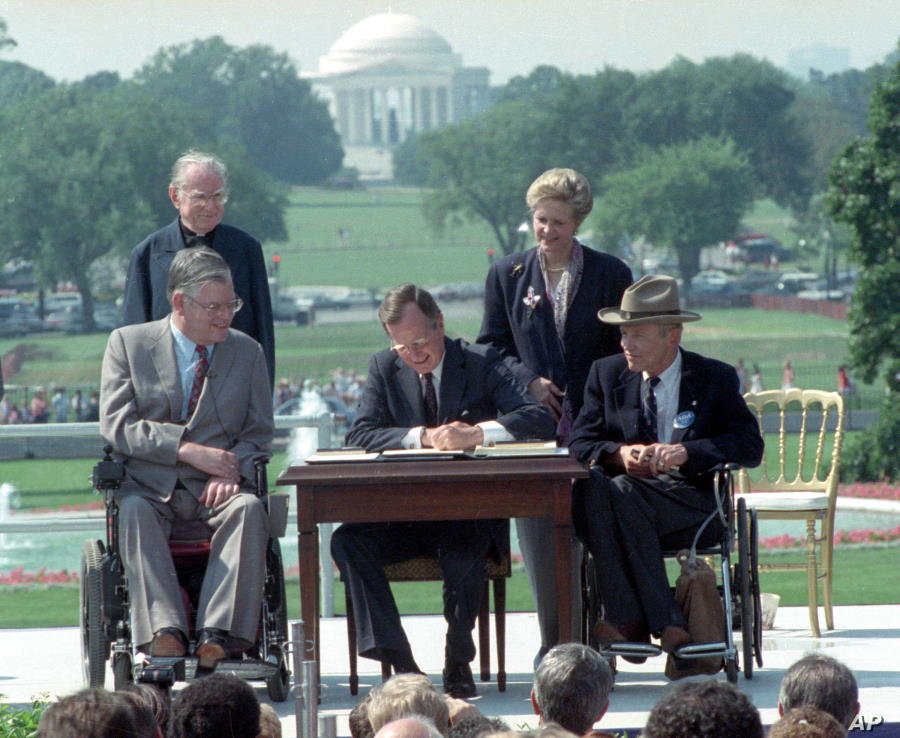 The president, flanked by two men in wheelchairs, with a man and woman standing behind him, signs a document at a small table. The backdrop is the U.S. Capitol Dome; it is a bright and sunny day on the Mall, and a crowd is gathered to watch.