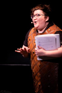 A performer holding a script during a reading, leaning forward to make a comic point. They are wearing glasses and a sort of brocaded orange cloak.