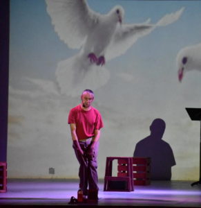 A production image with a man in a red shirt and walker standing in front of a projection of a white bird in flight.