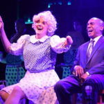 A woman in period clothes and a blonde wig (shie's playing Roxie Hart) sits and sings with a laughing gentleman in a suit (he's playing Billy Flynn). We're pretty sure this is the song "We Both Reached for the Gun."