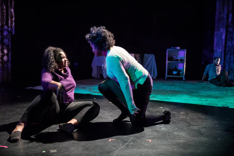 Women in a starkly lit, dramatic scene, one sprawled on the floor listening to another kneeling at her side.