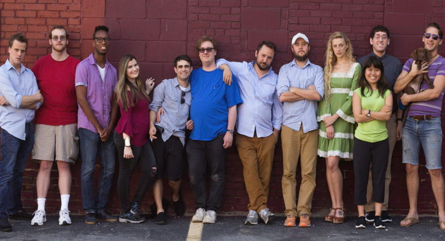 A dozen members of the theatre troupe (one holding a cute little brown dog) stand against a red brick wall.