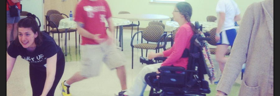 A lively moment in a classroom/rehersal setting; a woman bends at the waist, looks off left, a young man at center is blurred in sudden motion, and a young woman in a wheelchair looks on.